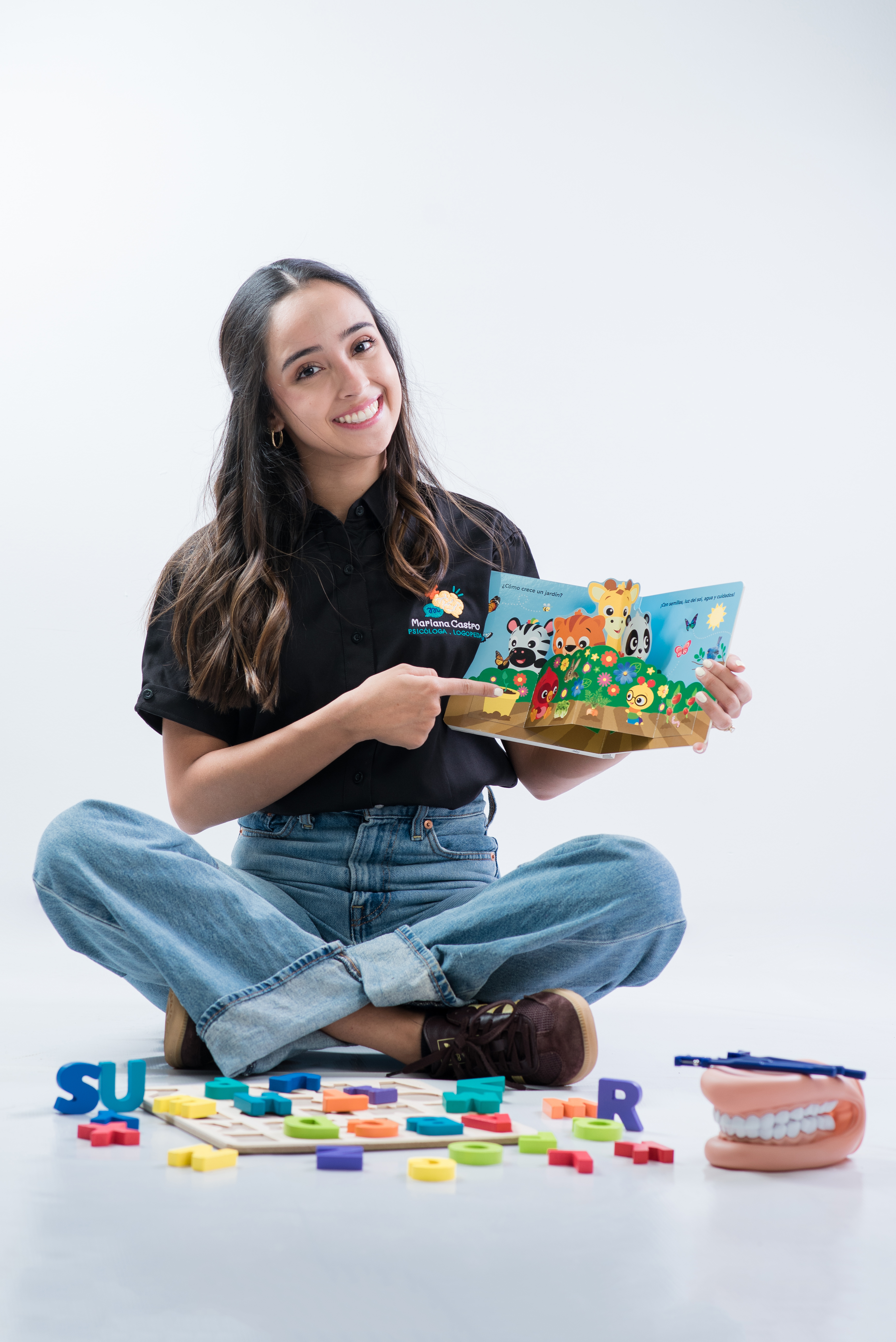 Mariana sentada en el piso con un libro y materiales educativos, mirando a la cámara con una sonrisa